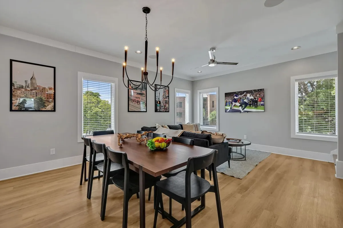 An inviting open-plan living and dining area featuring a dining table, chandelier, comfortable sofa, television, and artistic wall décor. The setting is lively and contemporary, perfect for both family gatherings and relaxed evenings.