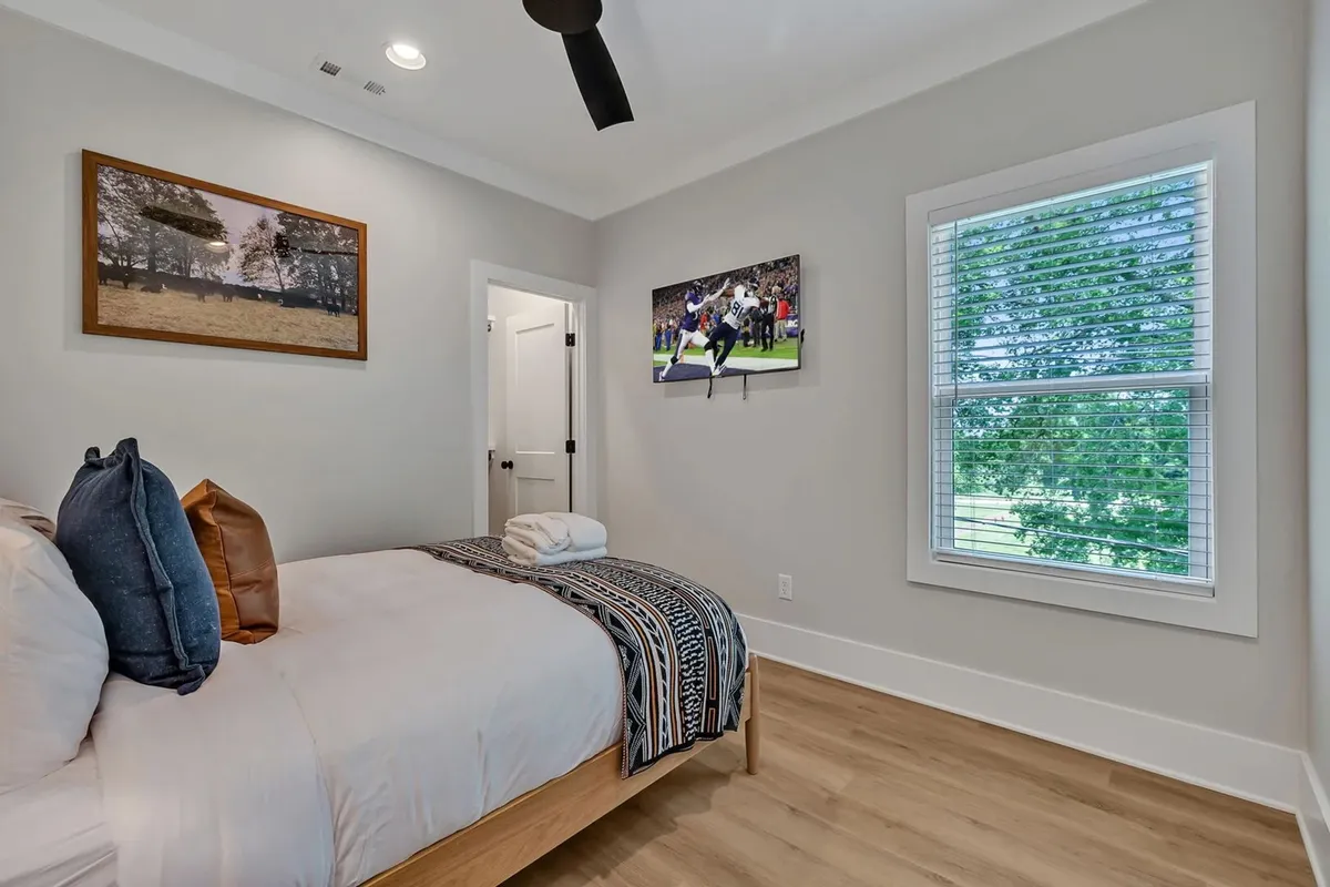 A stylish primary suite featuring a king bed dressed in plush linens, a smart TV mounted opposite the bed displaying a lively sports scene, framed artwork on the wall, and a large window framing lush greenery. The mood is cozy and inviting, making it perfect for relaxation and unwinding.