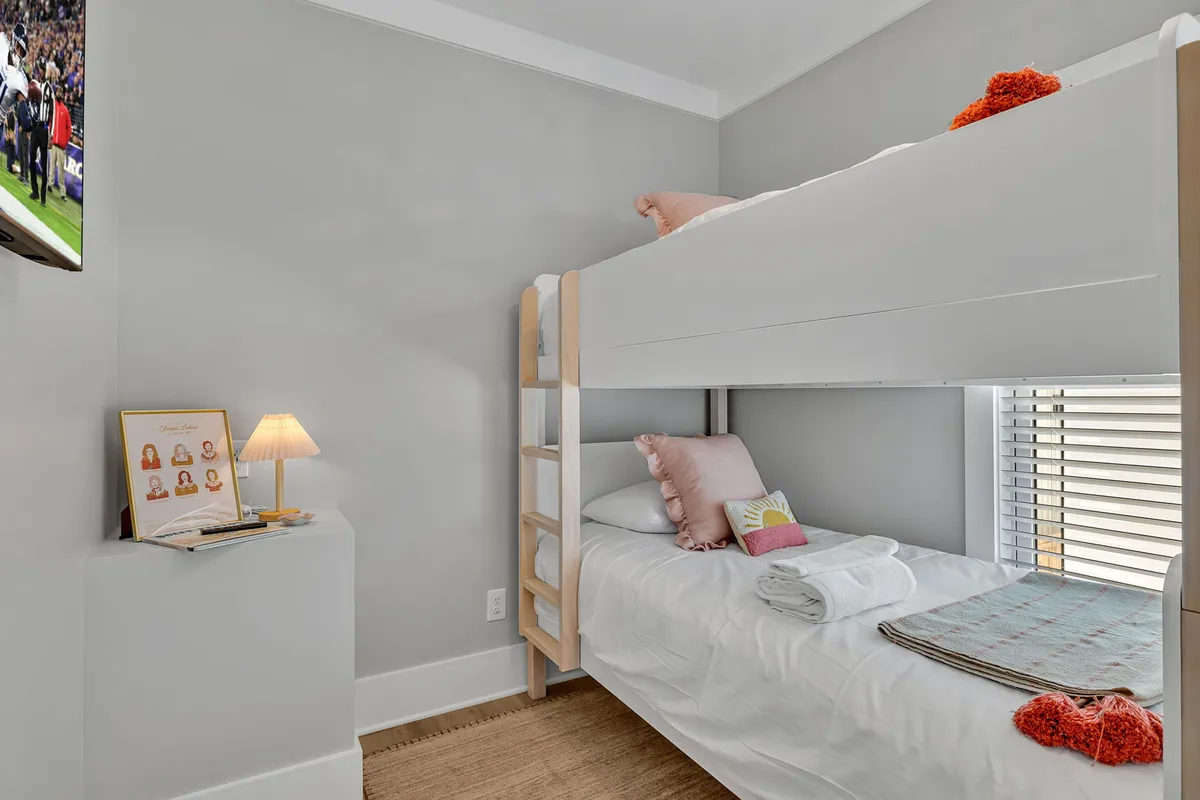 A playful and comfortable bunk room with twin beds, each adorned with colored pillows, a bedside lamp, framed photographs, and a wall-mounted television. The scene is warm and suited for shared evenings, suggesting versatility for both kids and extra guests.