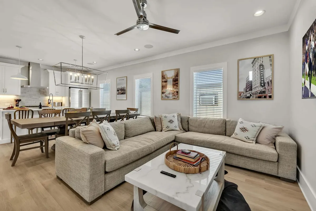 This image captures a spacious open-plan living and dining area, featuring a large sectional sofa, a generous dining table, decorative wall art, and a modern ceiling fan. The arrangement encourages group relaxation and social gatherings, with sightlines between the living and dining zones.