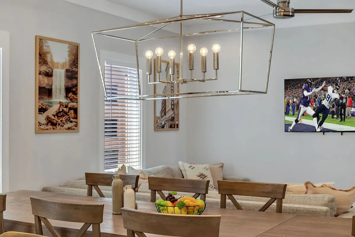 A warm dining area with a chandelier, a large dining table set for eight, bright wall art, and a television in the background. The space is designed for special occasions, group meals, and casual entertainment, blending style with inviting functionality.