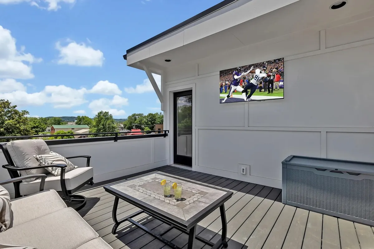 This image shows a private rooftop deck with a comfortable outdoor seating area, a wall-mounted TV, a coffee table set with drinks, and an expansive view of the surrounding landscape. The open-air setting is designed for relaxation and social gatherings, capturing the essence of a rooftop retreat.